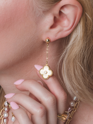 A woman’s ear is adorned with the Kinsey Designs Vivienne Quatrefoil Drop Earring in gold and white as her pink nails gently touch her chin.