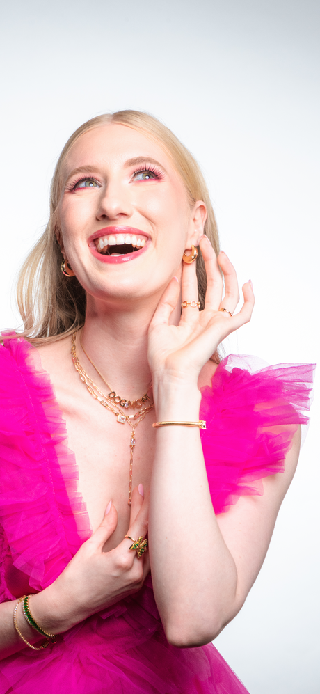 A smiling woman in a bright pink dress wears gold jewelry, featuring the stunning Kinsey Designs Archives Yara Lariat gold-filled cubic zirconia necklace, as she touches her face and looks upward.