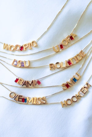 Kinsey Designs’ Stone Spacer Slides, available in 9 colors, accent gold-filled charm necklaces that spell out college team chants like “ROLL TIDE” and “HOOK EM,” all styled on a crisp white background.