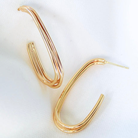 The Dutton Earring by Kinsey Designs Archives features gold-plated, oval-shaped hoops with a layered design, measuring 1.5" long and 0.6" wide, shown on a white fabric background.