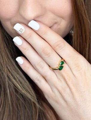 A woman with white nail polish and a Kinsey Designs Archives Teara Ring - Emerald, adorned with green stones and cubic zirconia, touches her lips, revealing a delicate flower nail design.