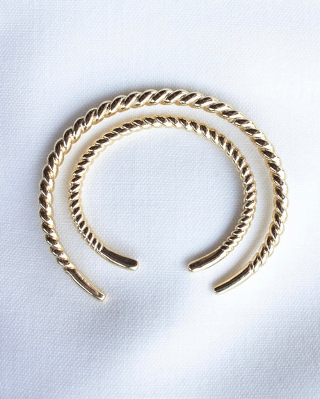 The Kinsey Designs Fallon Baby Cuff features two gold-filled twisted open bangles arranged concentrically on white textured fabric, creating a stylish and nickel-free adjustable bracelet.