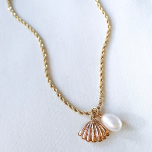 The Cove Pearl Necklace by Kinsey Designs Archives features a gold chain with a delicate seashell locket and white pearl pendant, elegantly showcased on a light background.