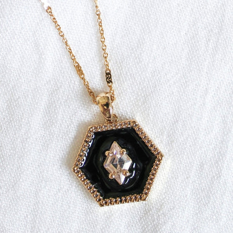 The Nova Necklace by Kinsey Designs Archives features a gold-filled chain with a hexagonal black pendant and a clear cubic zirconia crystal at the center, elegantly displayed on white fabric.