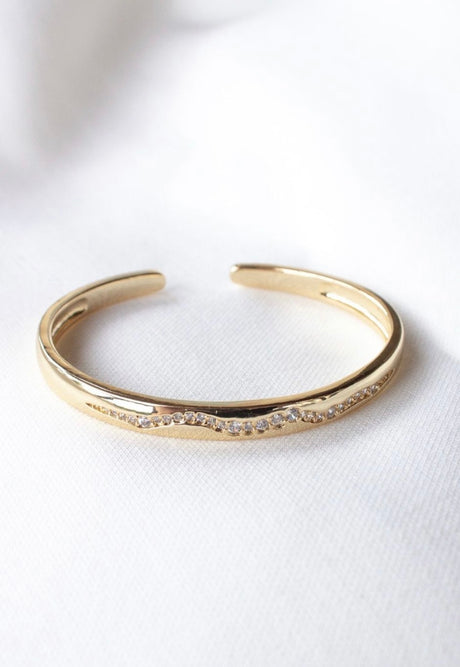 The Lily Cuff by Kinsey Designs Archives is a gold filled bracelet featuring a row of sparkling cubic zirconia stones on a white fabric background. This adjustable cuff brings elegant style to any outfit.