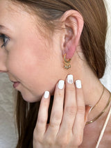 A woman wearing a 1-inch long gold filled Tove Huggie by Kinsey Designs Archives, with a white manicure and layered gold necklaces, touches her face.