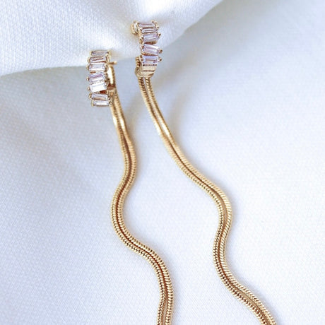 The Callie Earring by Kinsey Designs Archives features gold filled baguette-cut cubic zirconia and long, wavy 3.5-inch snake chain drops, displayed on white fabric.