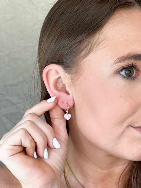 A woman with white nails touches her ear, showcasing the Kinsey Designs Archives Cutie Huggie—gold filled, heart-shaped earrings. These elegant 1-inch huggies glimmer beautifully with every movement.