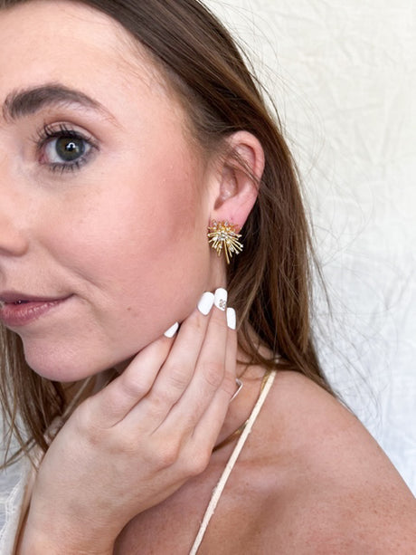 A woman with brown hair touches her ear, showing off Kinsey Designs Archives’ Harlem Post gold-plated burst-shaped earrings accented with cubic zirconia and manicured white nails.