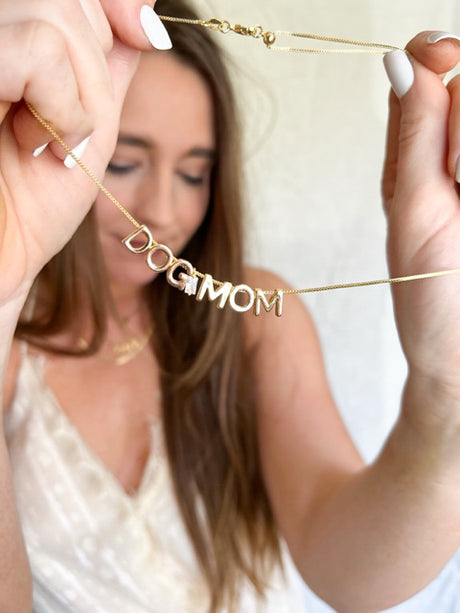 A woman holds the Kinsey Designs Archives Dog Mom Slide Necklace, featuring gold filled letters spelling "DOG MOM" on an adjustable Italian chain.