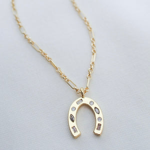 The Delphin Necklace by Kinsey Designs features a gold filled horseshoe pendant adorned with small gemstones, symbolizing luck. This shimmering luck necklace is shown on a white background.