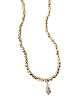 The Penn Necklace by Kinsey Designs Archives is a gold beaded necklace featuring a clear, pear-shaped crystal pendant, shown on a white background.