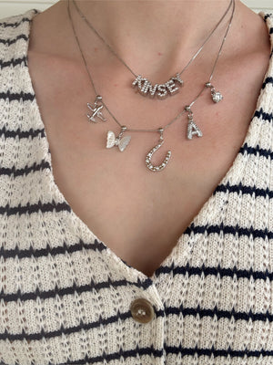 Close-up of three layered Kinsey Designs Charm Bar Necklaces featuring Silver Birthstone Grip Charms, styled over a cream knit top with black stripes and a button.