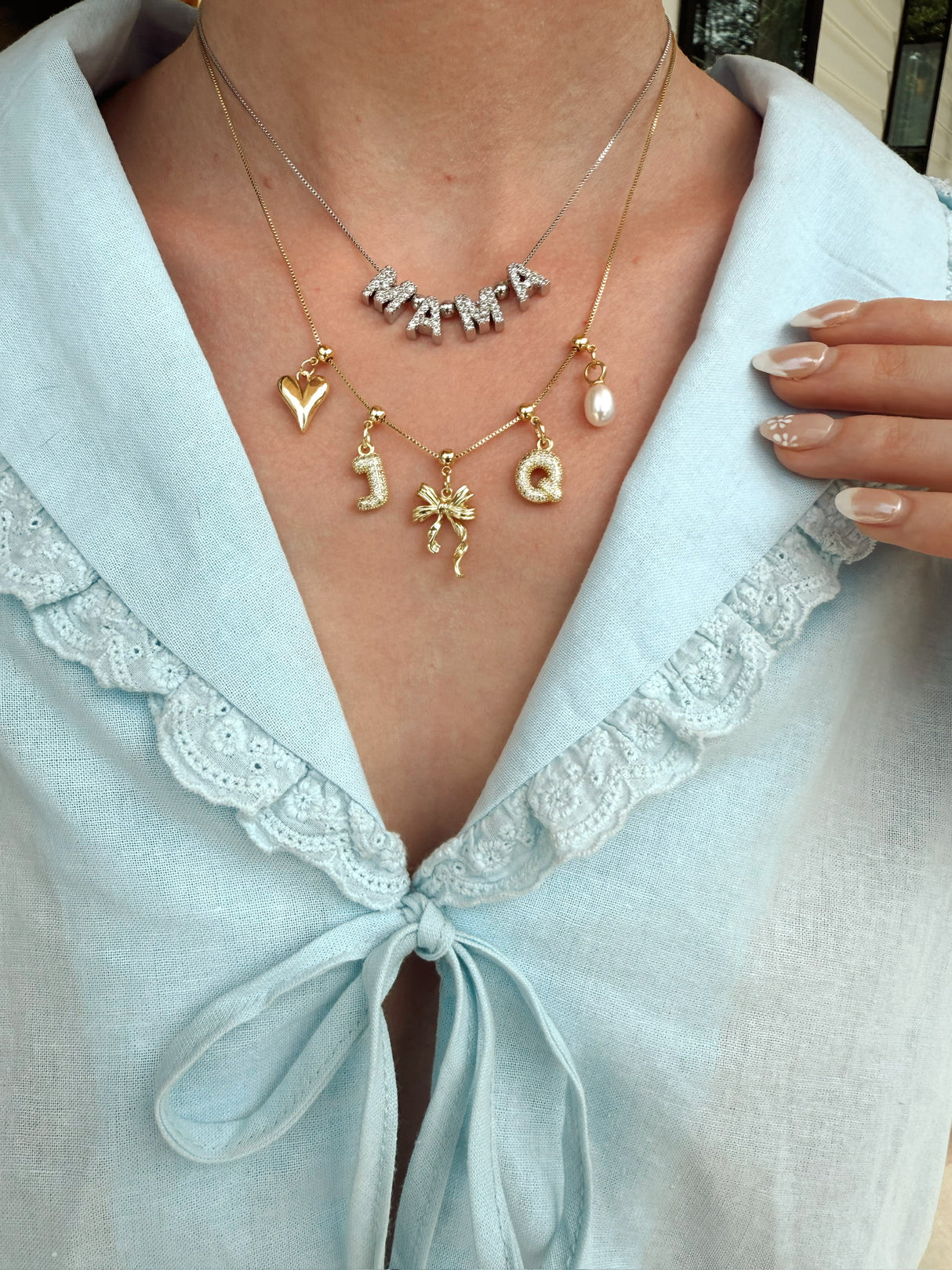 A woman wears three Kinsey Designs Round Spacer Slide necklaces, each featuring a delicate charm, paired with a light blue lace-trim blouse and bow.