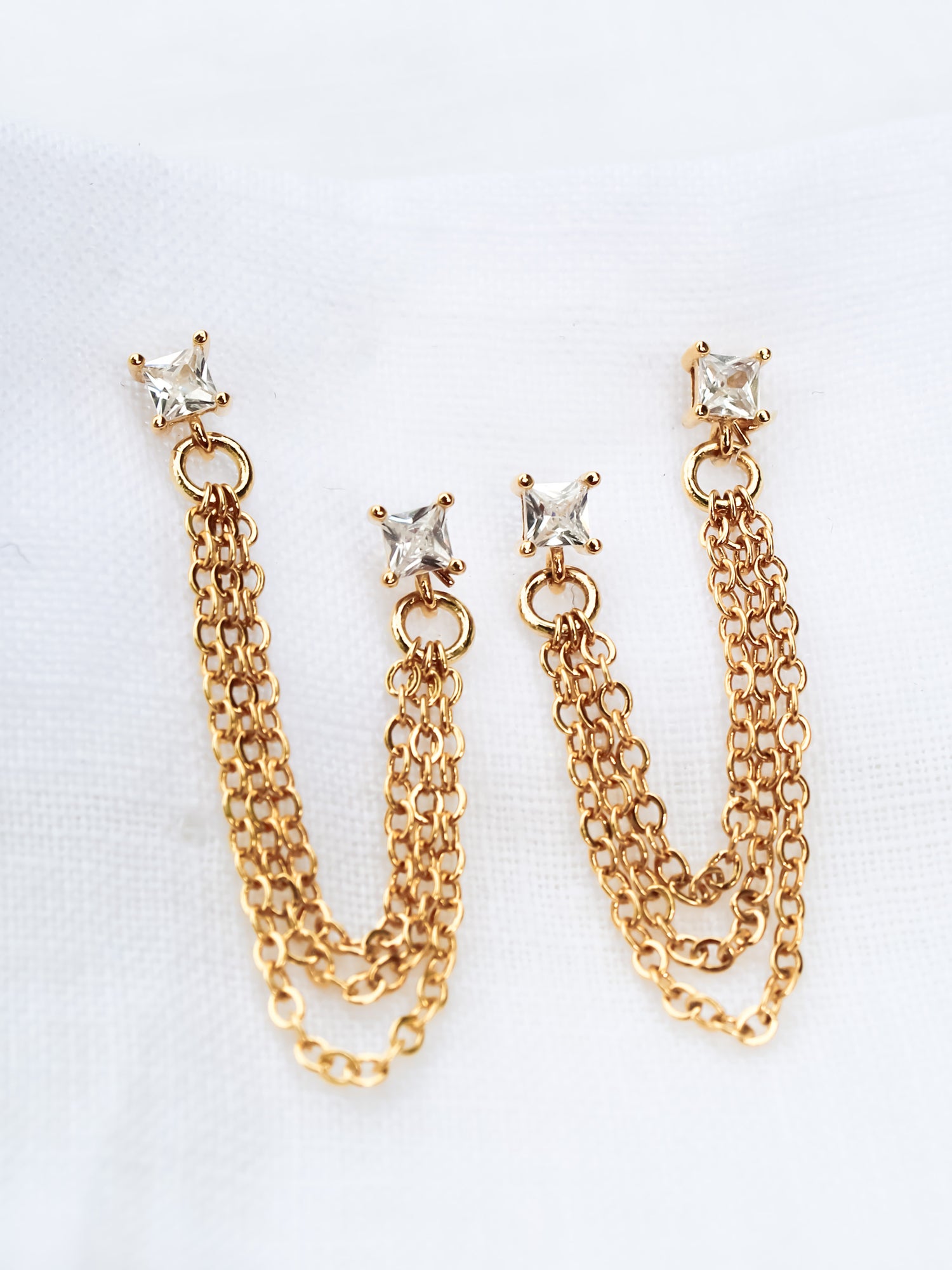 The Becca Double Post by Kinsey Designs Archives features gold-plated chain earrings with square crystal studs, measuring 2" long from post to post, displayed on a white fabric background.