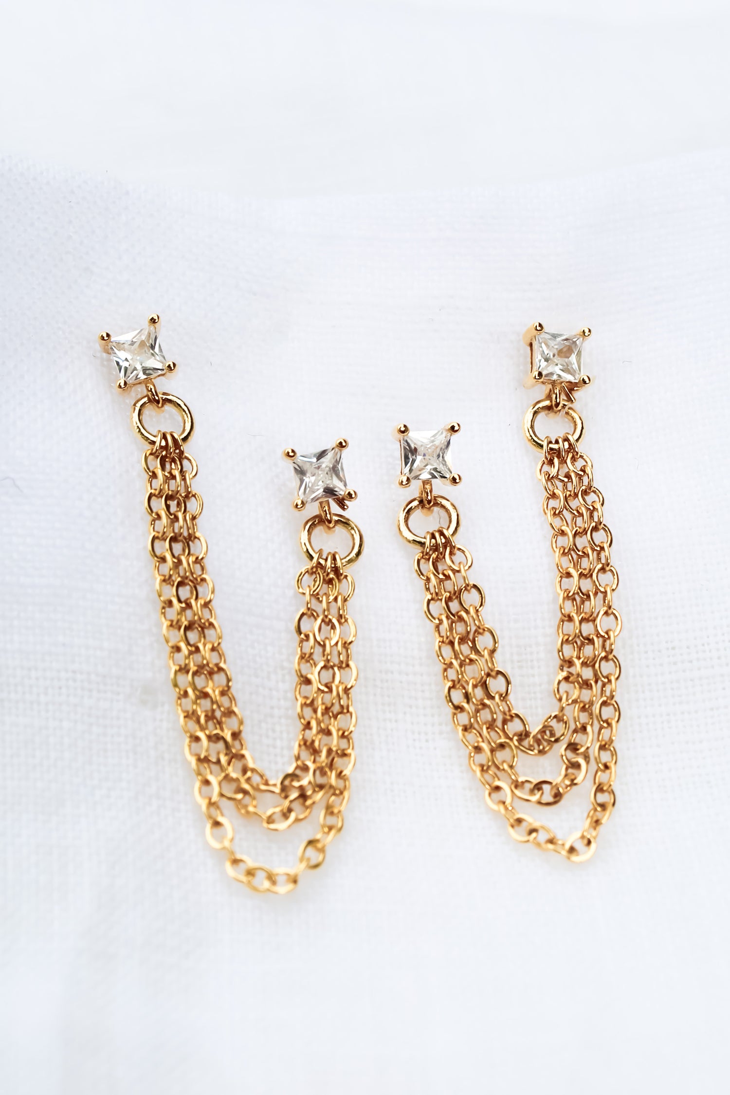 The Becca Double Post by Kinsey Designs Archives features gold-plated chain earrings with square crystal studs, measuring 2" long from post to post, displayed on a white fabric background.