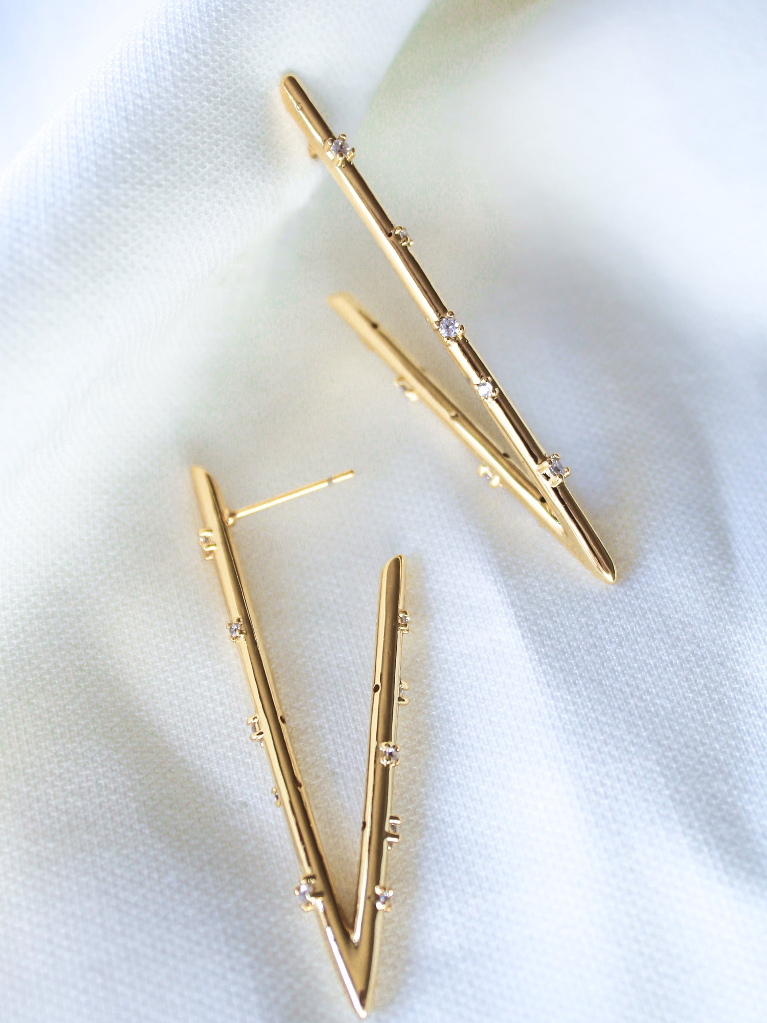 The Bowen Earring Luxe by Kinsey Designs features gold filled V-shaped earrings adorned with small cubic zirconia gemstones, shown on a soft white textured fabric background.
