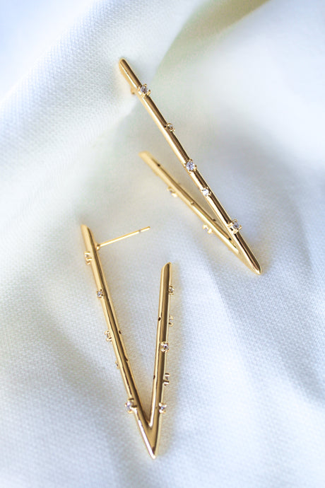 The Bowen Earring Luxe by Kinsey Designs features gold filled V-shaped earrings adorned with small cubic zirconia gemstones, shown on a soft white textured fabric background.