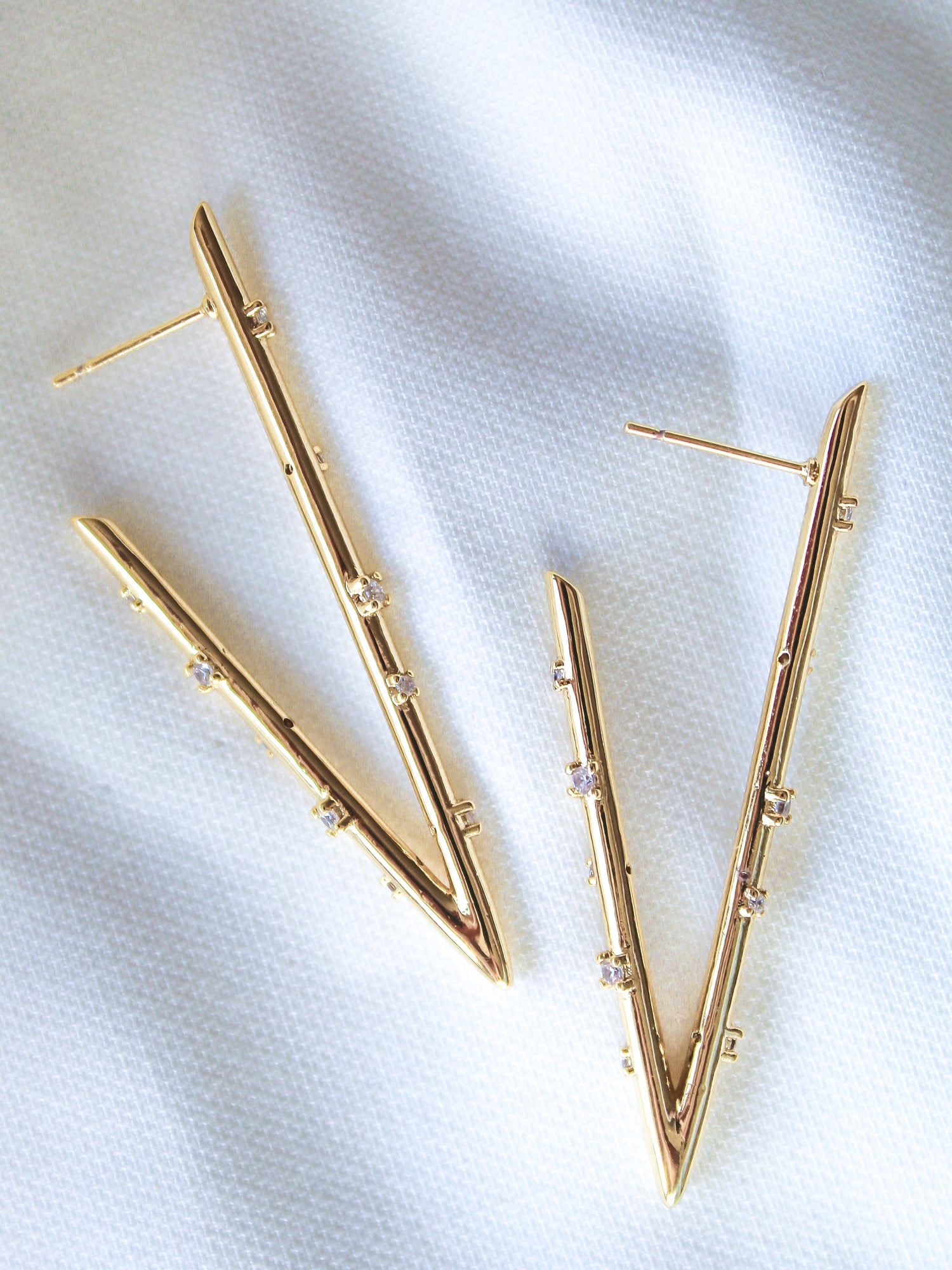 The Bowen Earring Luxe by Kinsey Designs features gold-filled V-shaped earrings with small cubic zirconia gemstones, shown on a white textured fabric background.