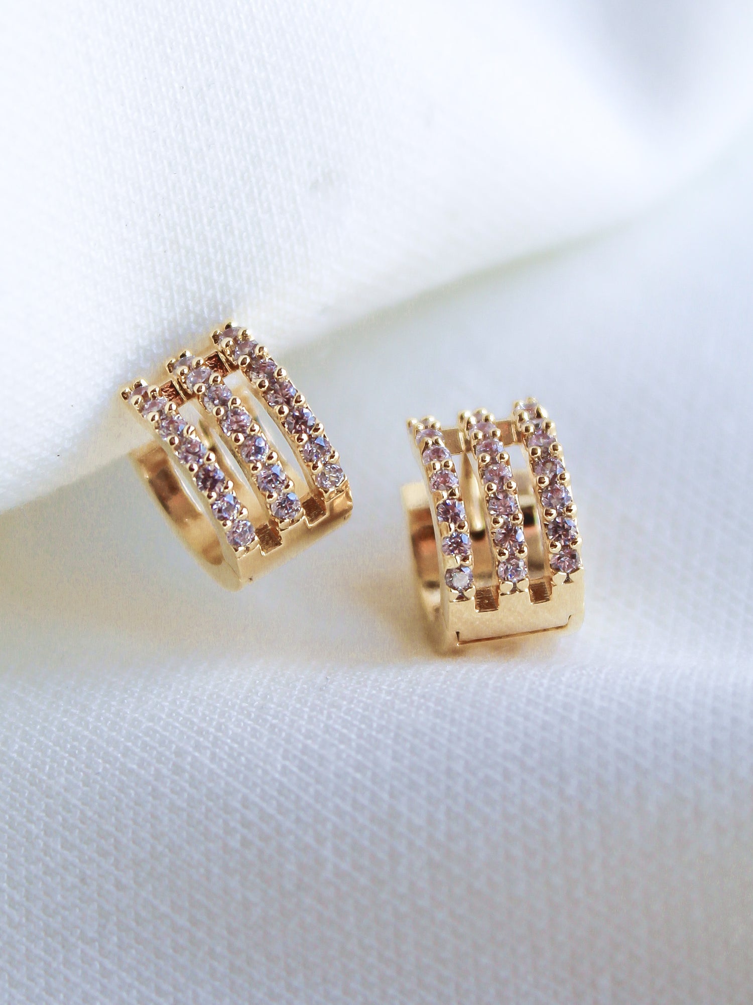 The Bri Huggie by Kinsey Designs Archives features 14k gold plating and three rows of sparkling cubic zirconia, shown against a white fabric background.