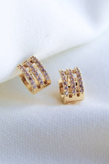 The Bri Huggie by Kinsey Designs Archives features 14k gold plating and three rows of sparkling cubic zirconia, shown against a white fabric background.