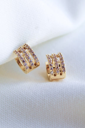 The Bri Huggie by Kinsey Designs Archives features 14k gold plating and three rows of sparkling cubic zirconia, shown against a white fabric background.