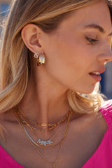 A woman in a pink top is shown in profile wearing Kinsey Designs Archives' Bri Huggie 14k gold plated earrings and layered gold necklaces.