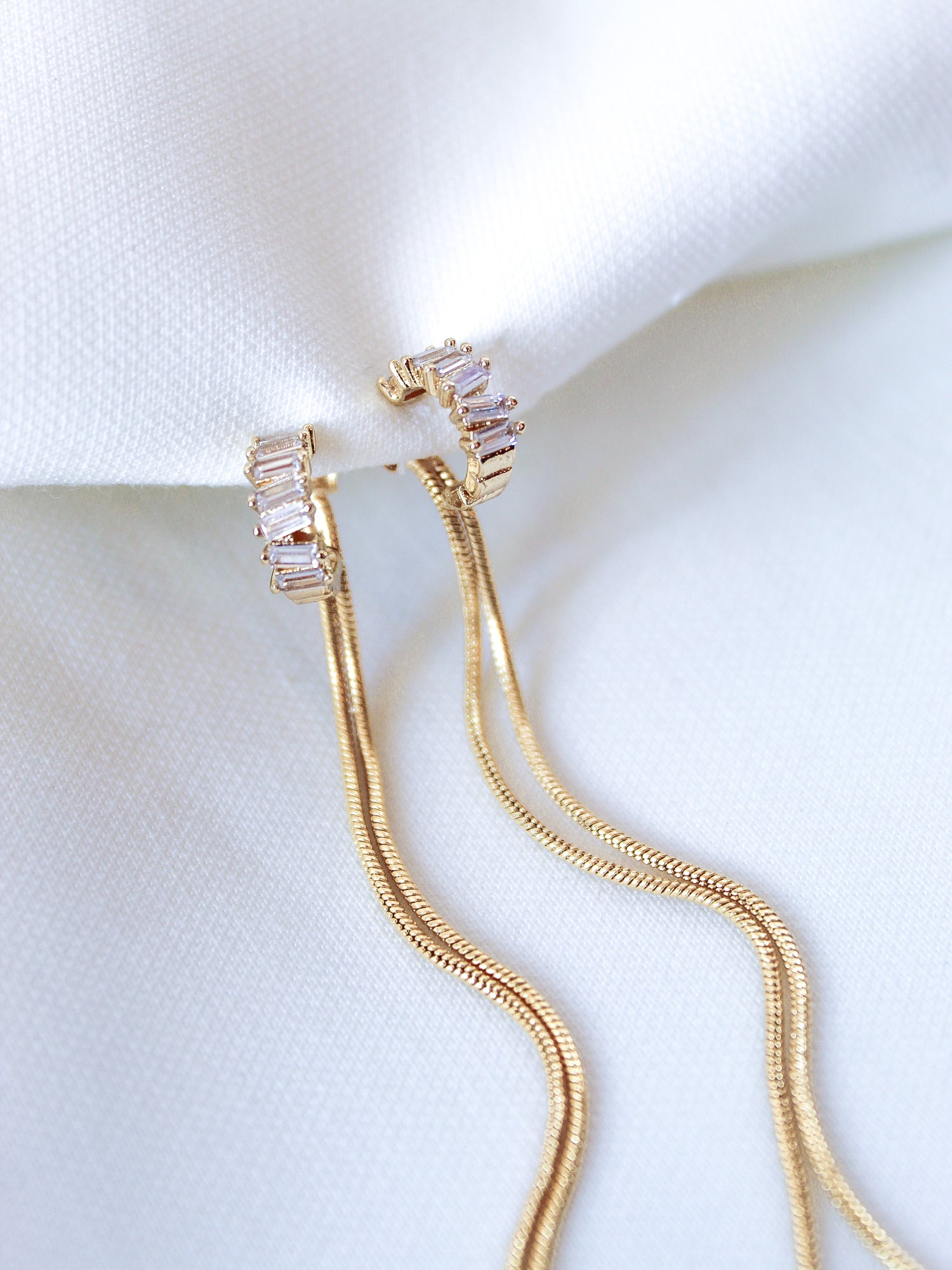 The Callie Earring by Kinsey Designs Archives features 3.5-inch gold-filled earrings with sparkling cubic zirconia drops and long chains, displayed on white textured fabric.
