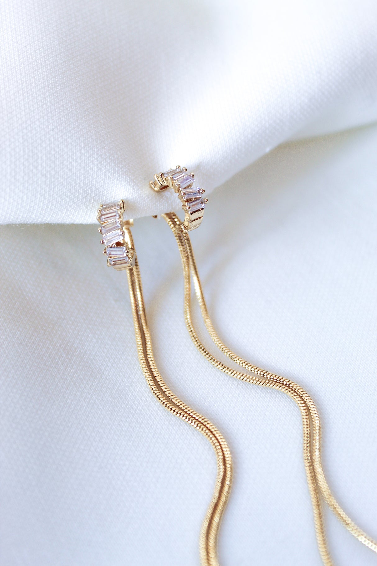 The Callie Earring by Kinsey Designs Archives features 3.5-inch gold-filled earrings with sparkling cubic zirconia drops and long chains, displayed on white textured fabric.