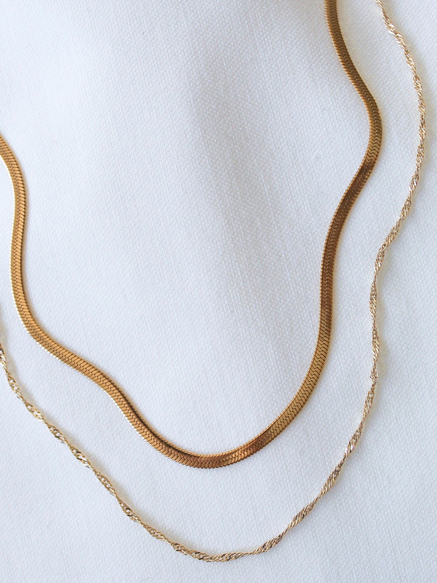 The Callum Layer by Kinsey Designs Archives features two 14k gold-filled necklaces—one flat, one twisted—styled as a layered necklace on white fabric. Both include a 3" extender for adjustable length.