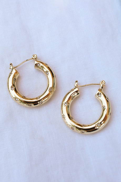 The Camber Earring by Kinsey Designs Archives is a 14k gold filled hoop with small engraved stars and a 1.25-inch circumference, shown on a white fabric background.