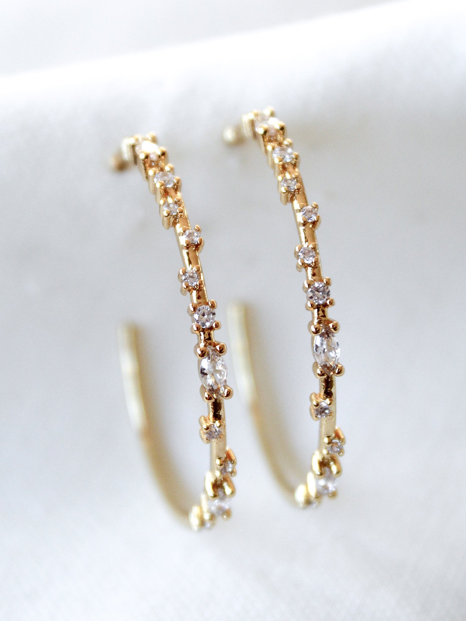 The Chase Hoop Large earrings by Kinsey Designs, gold filled and embellished with sparkling gemstones, are displayed on a white fabric background.