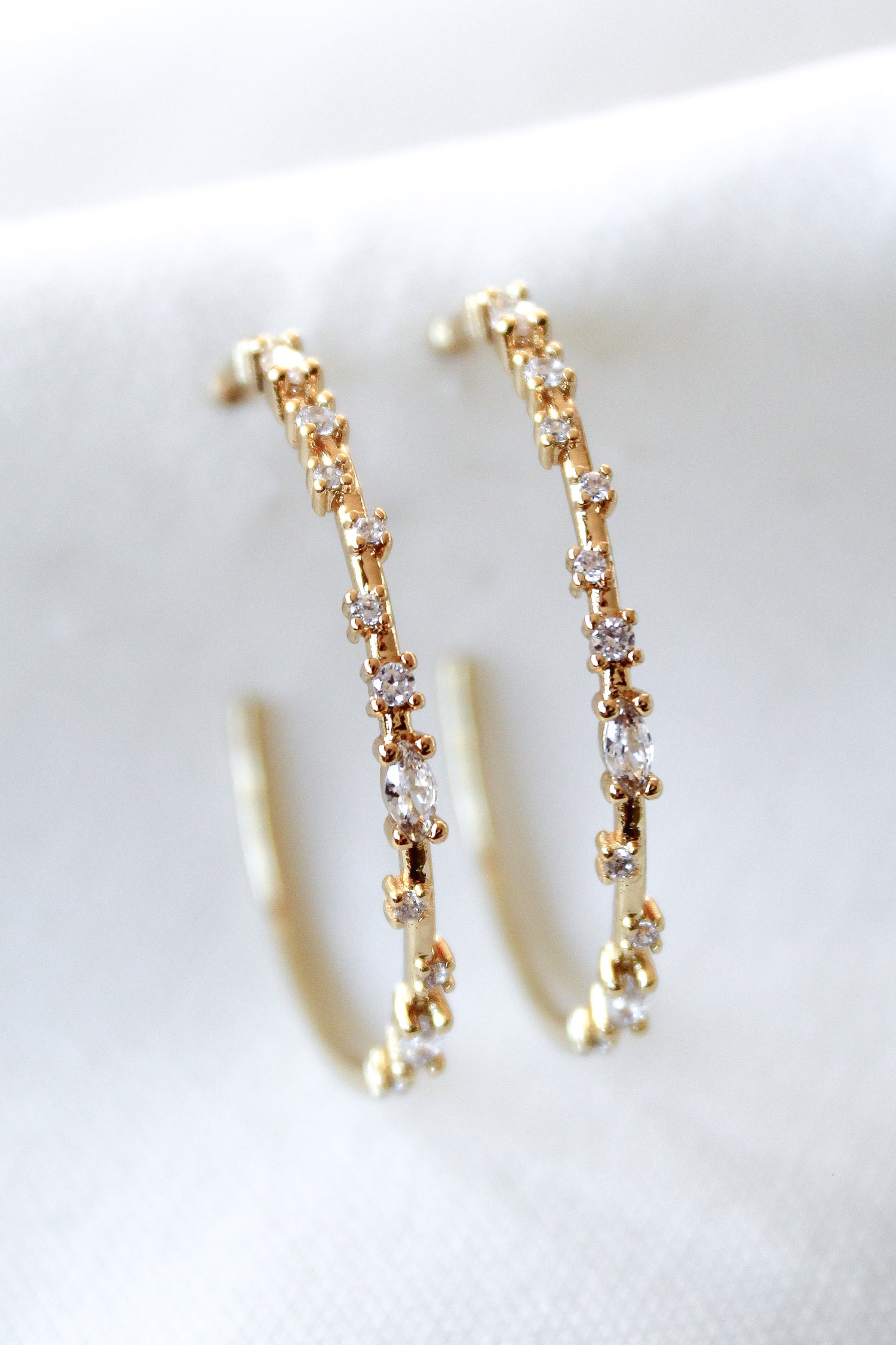 The Chase Hoop Large earrings by Kinsey Designs, gold filled and embellished with sparkling gemstones, are displayed on a white fabric background.