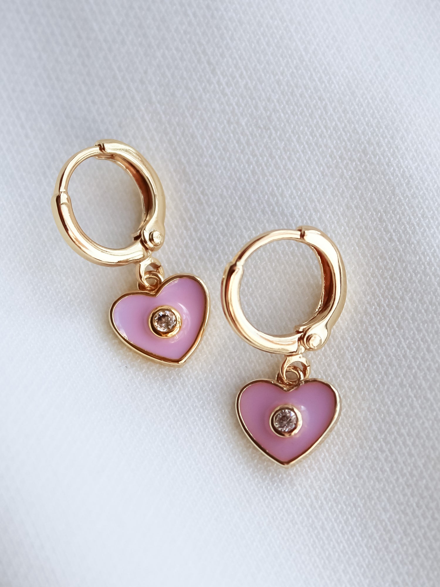The Cutie Huggie by Kinsey Designs Archives: 1-inch gold-filled earrings with pink heart-shaped charms and a central gemstone, displayed on white fabric.