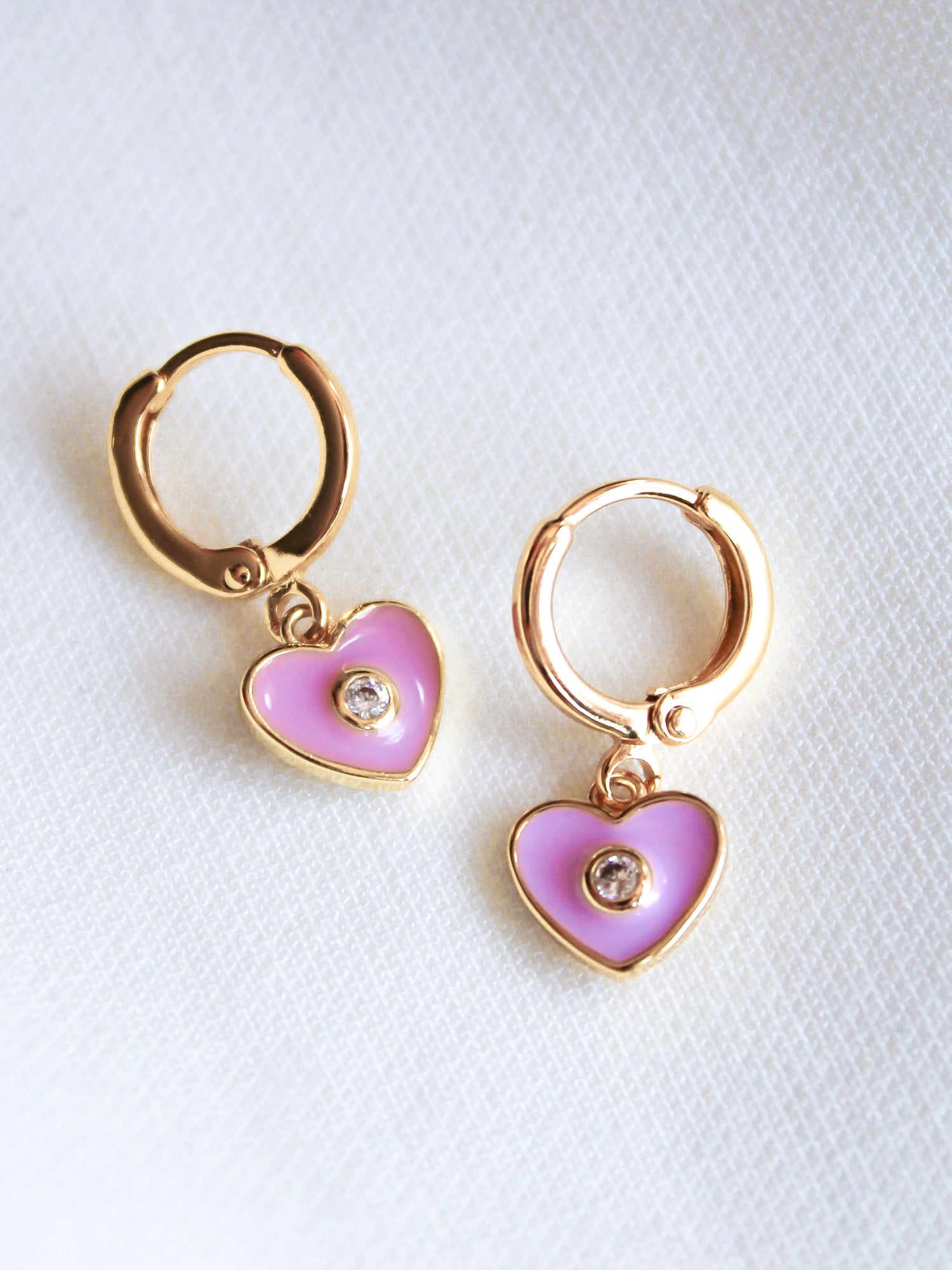 The Cutie Huggie by Kinsey Designs Archives features 1-inch gold filled earrings with pink heart-shaped charms and a central gem, shown on a light fabric background.