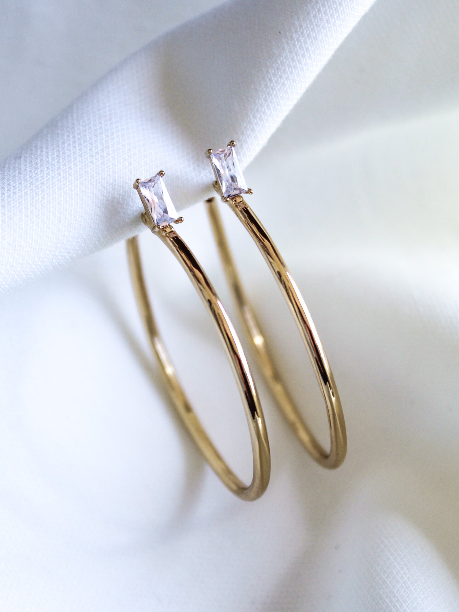The Darra Hoop Large by Kinsey Designs Archives features gold-plated hoops adorned with rectangular cubic zirconia, shown on a white fabric background.