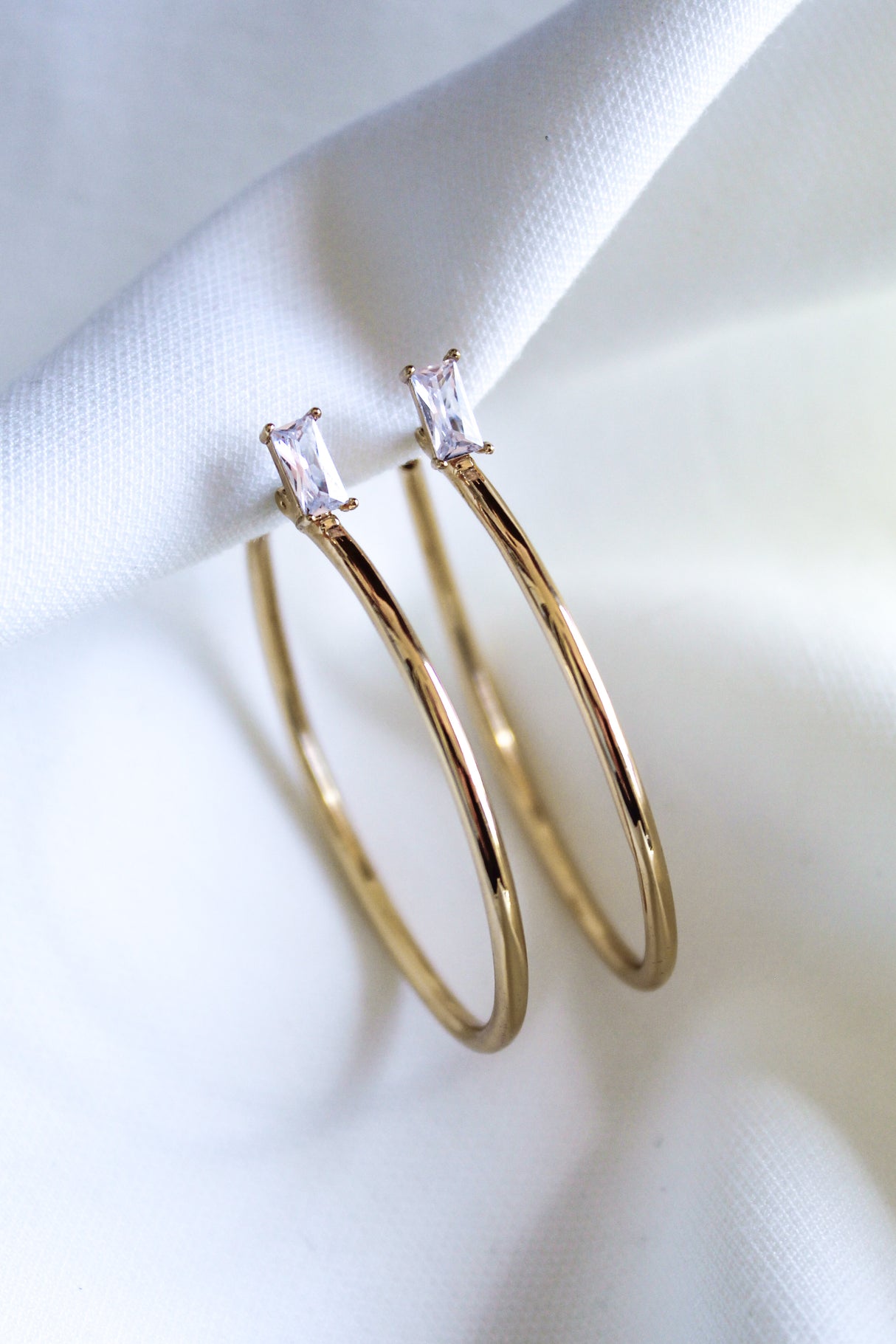 The Darra Hoop Large by Kinsey Designs Archives features gold-plated hoops adorned with rectangular cubic zirconia, shown on a white fabric background.