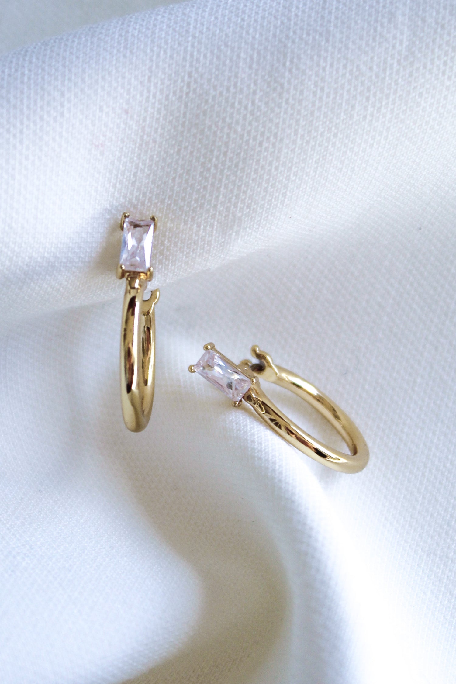 The Darra Hoop Mini by Kinsey Designs Archives are gold-plated hoops with a 0.75" diameter, featuring rectangular clear cubic zirconia stones displayed on soft white fabric.