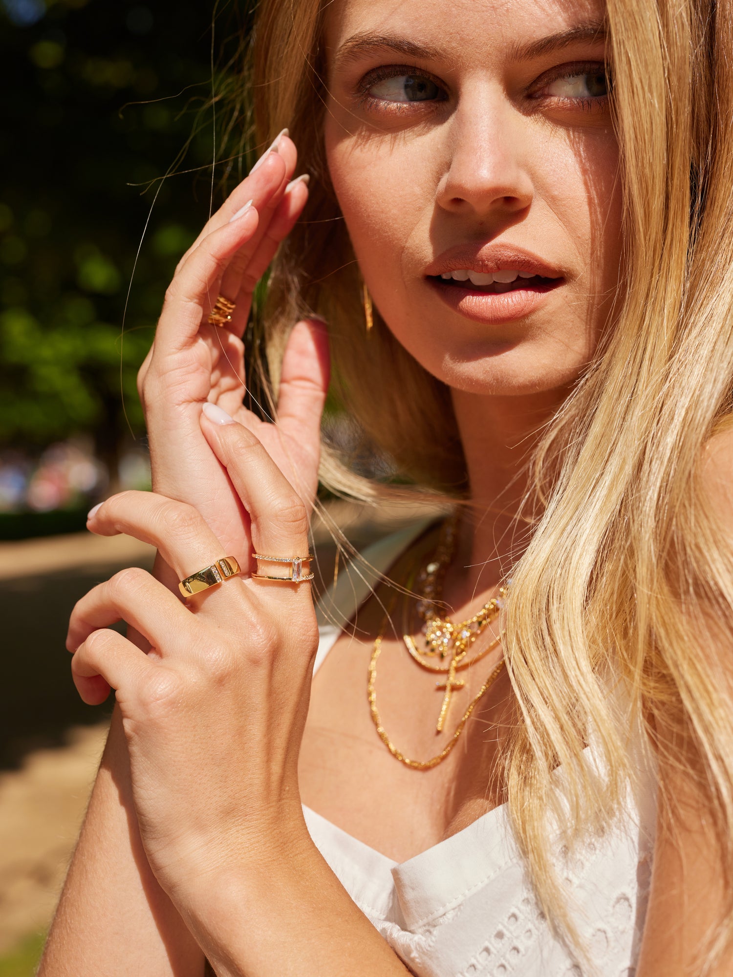 A woman in sunlight, wearing a white dress, touches her face and looks to the side while showcasing the Kinsey Designs Archives Dean Ring, a gold-plated cubic zirconia ring in size 6.