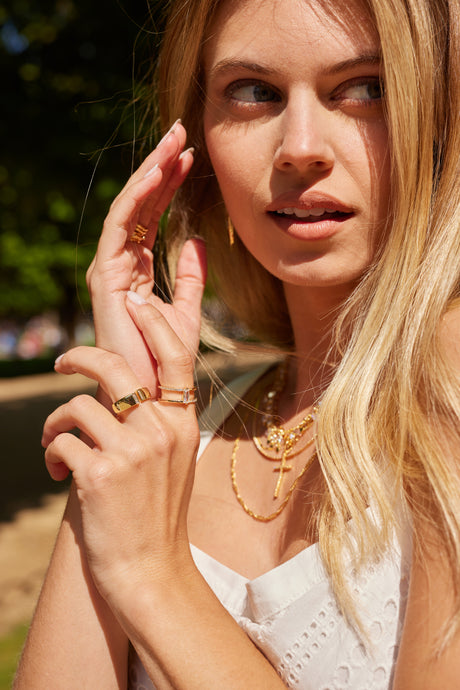 A woman in sunlight, wearing a white dress, touches her face and looks to the side while showcasing the Kinsey Designs Archives Dean Ring, a gold-plated cubic zirconia ring in size 6.