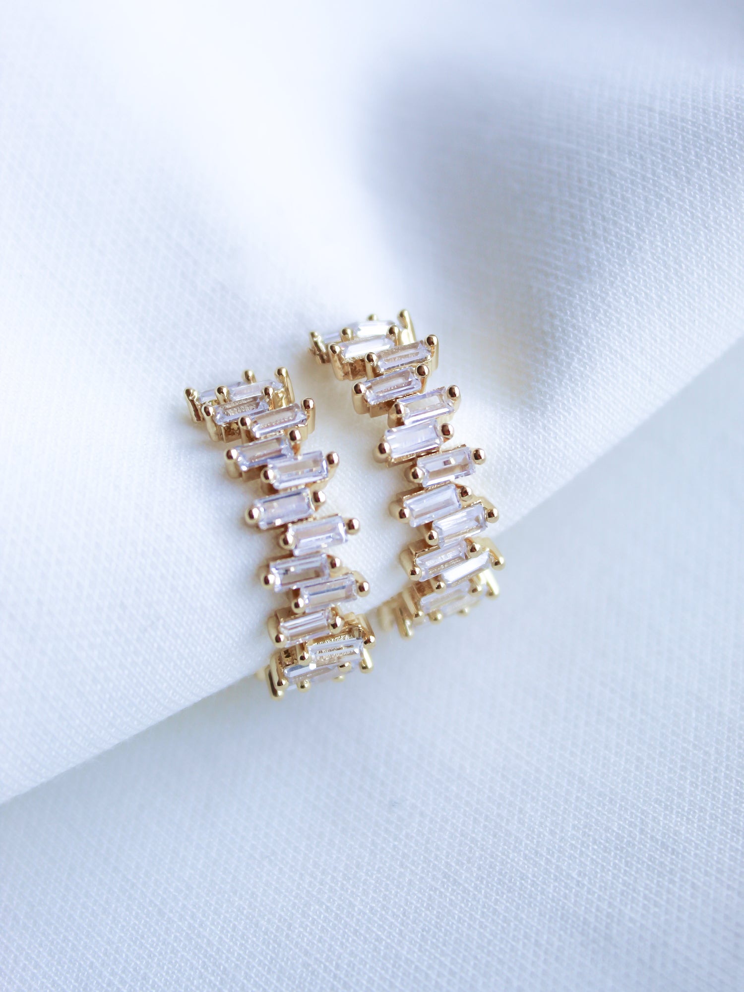 The Ettie Hoop by Kinsey Designs Archives are gold-filled earrings with a 1" diameter, featuring rectangular cubic zirconia stones in a zigzag pattern, shown on white fabric.