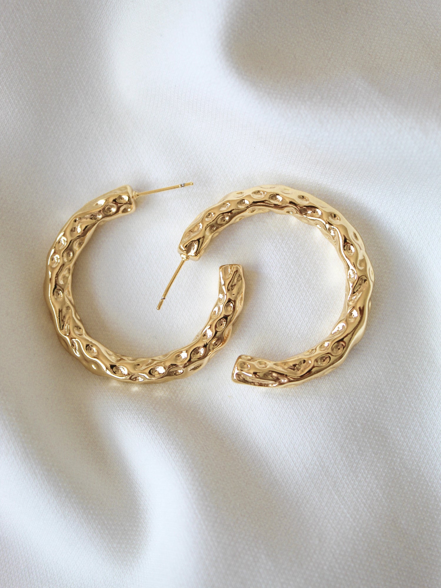 The Hammer Hoop earrings by Kinsey Designs Archives, featuring a textured gold-filled design and a 1.5" diameter, are displayed on soft white fabric to showcase their elegant style.