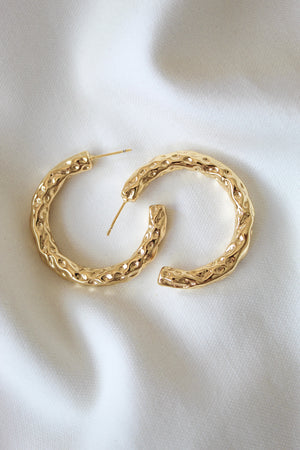 The Hammer Hoop earrings by Kinsey Designs Archives, featuring a textured gold-filled design and a 1.5" diameter, are displayed on soft white fabric to showcase their elegant style.