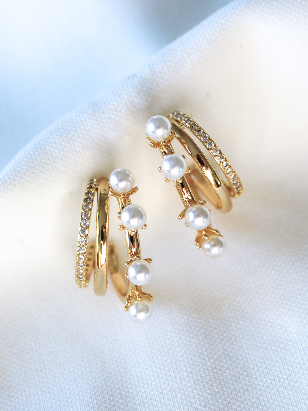 The Hattie Pearl Hoops by Kinsey Designs feature a triple-layer gold-filled design with pearl accents, adding a modern twist to classic hoops. Shown on a white fabric background.