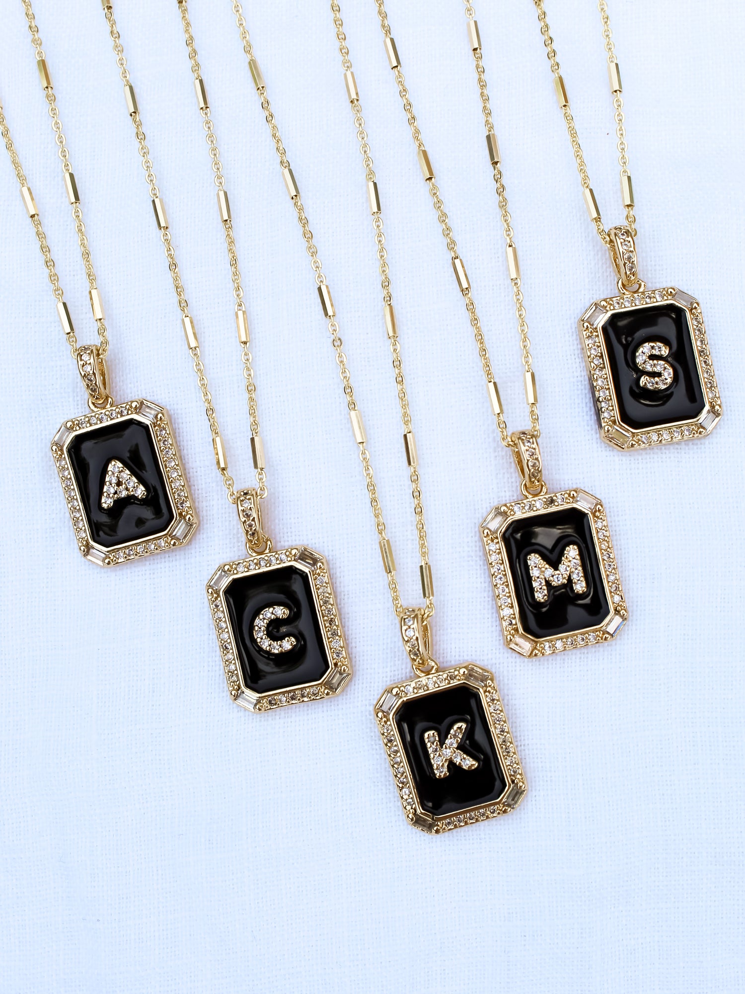 Five gold necklaces from Kinsey Designs Archives, each with a black pendant and a different silver letter—A, G, K, M, S—like the Initial Tile Necklace Ink Letter V.
