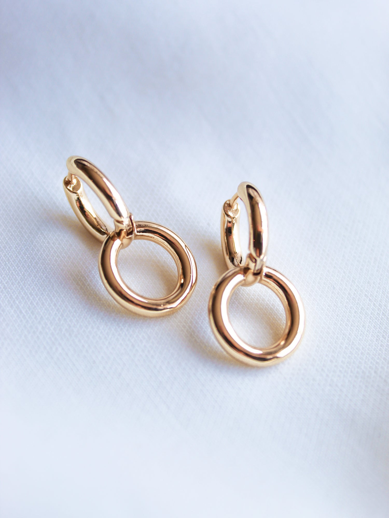 The Interlock Huggie earrings by Kinsey Designs Archives, featuring double 14k gold plated circles, are displayed on a white fabric background.