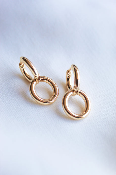 The Interlock Huggie earrings by Kinsey Designs Archives, featuring double 14k gold plated circles, are displayed on a white fabric background.