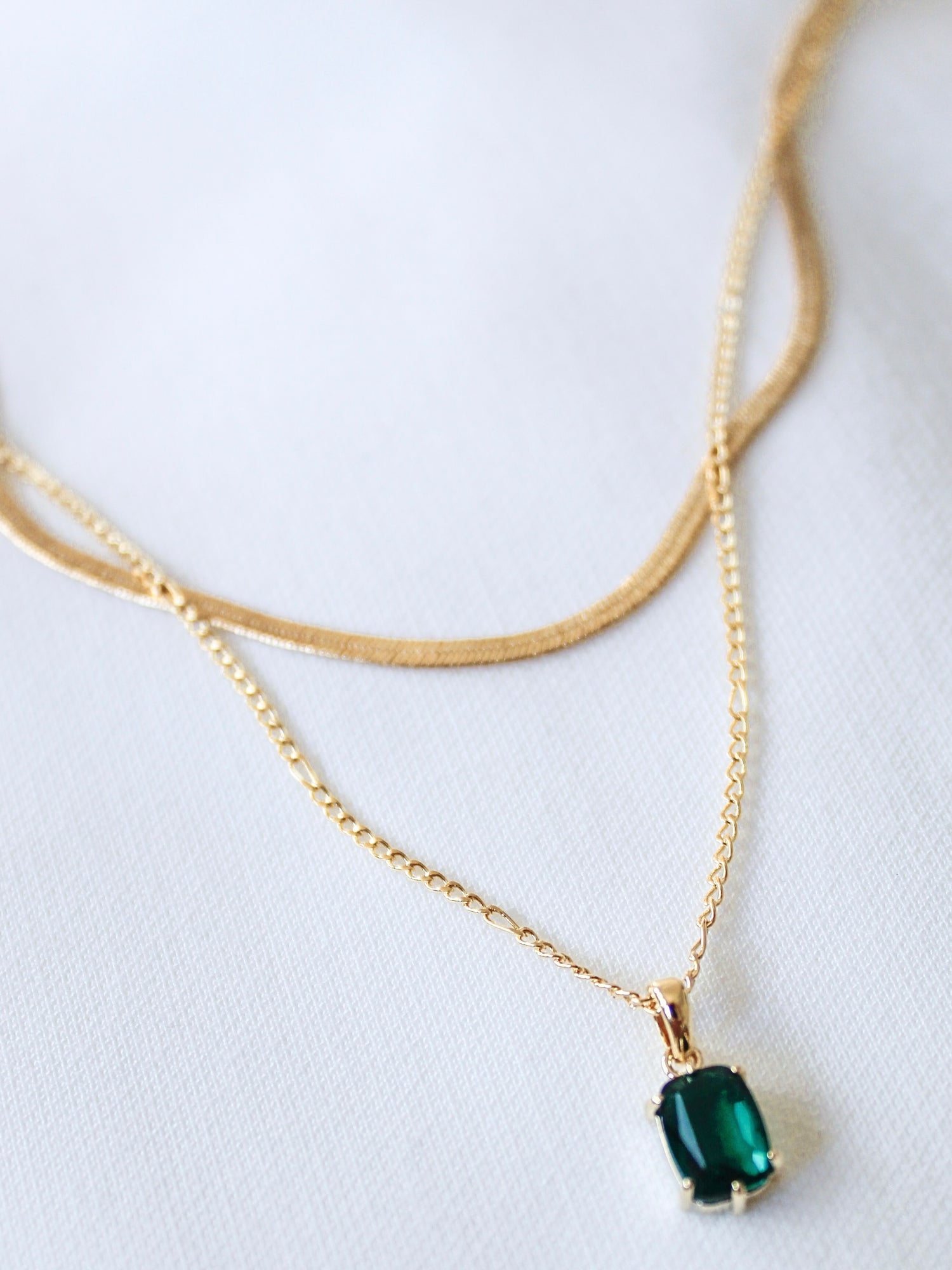 The Kelley Layer by Kinsey Designs Archives features an 18" gold plated chain with a bold green gemstone pendant, elegantly showcased on a white fabric background.