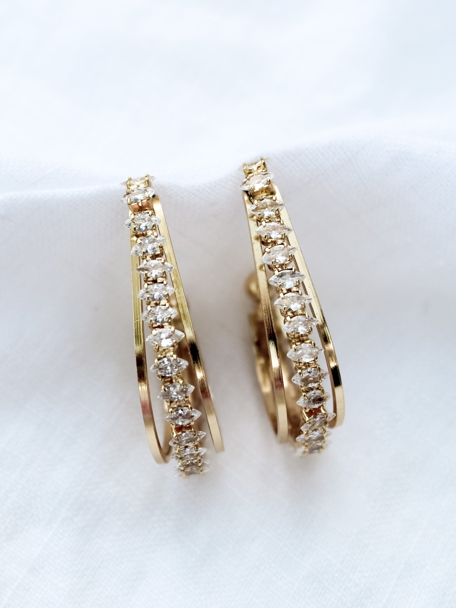The Kenny Earring by Kinsey Designs Archives features gold-filled hoops embellished with rows of sparkling cubic zirconia, elegantly presented on a white fabric background.