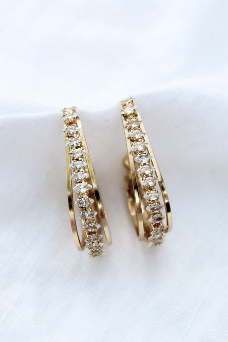 The Kenny Earring by Kinsey Designs Archives features gold-filled hoops embellished with rows of sparkling cubic zirconia, elegantly presented on a white fabric background.